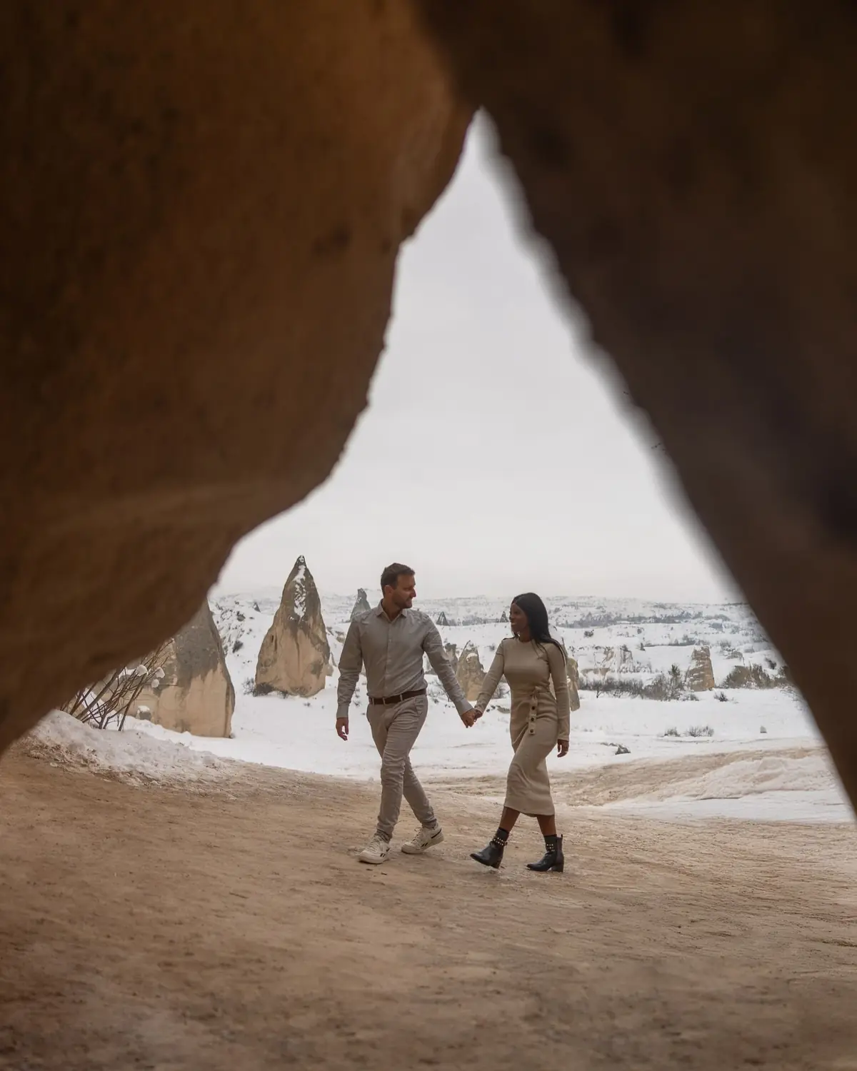 Us walking hand in hand, framed through a cave mouth, snowy fairy chimneys beyond