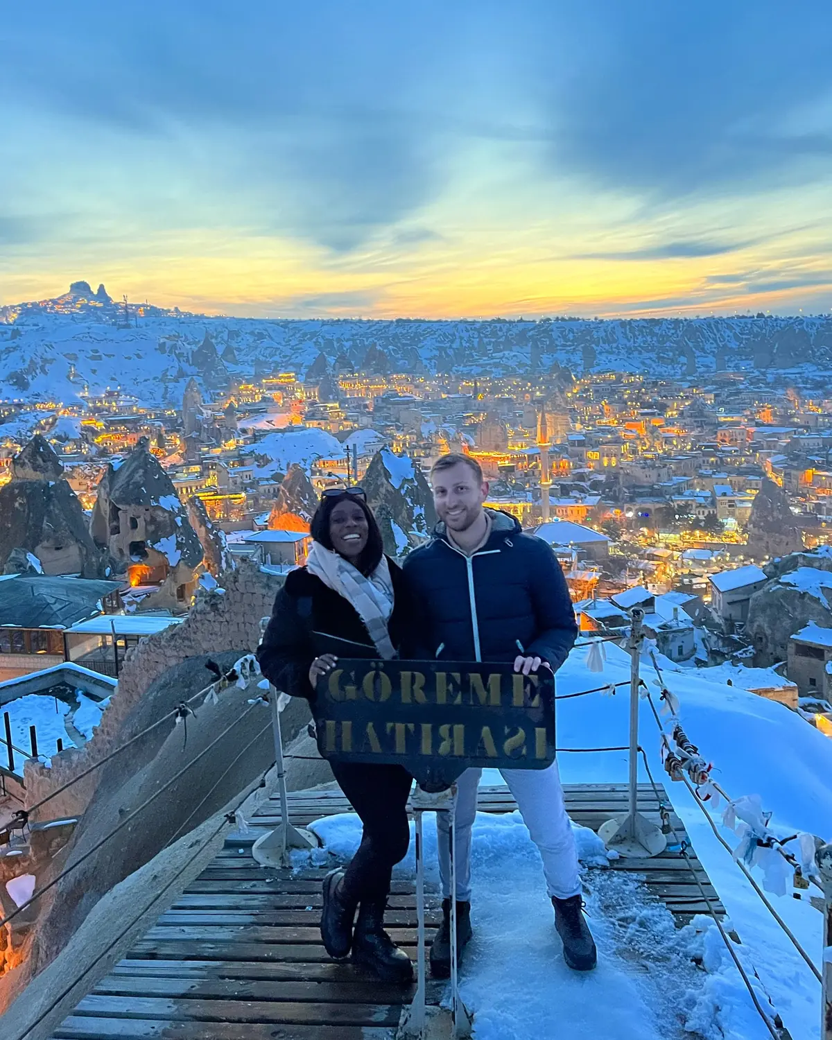 Us holding a Göreme sign at a sunset viewpoint over the twinkling snowy town