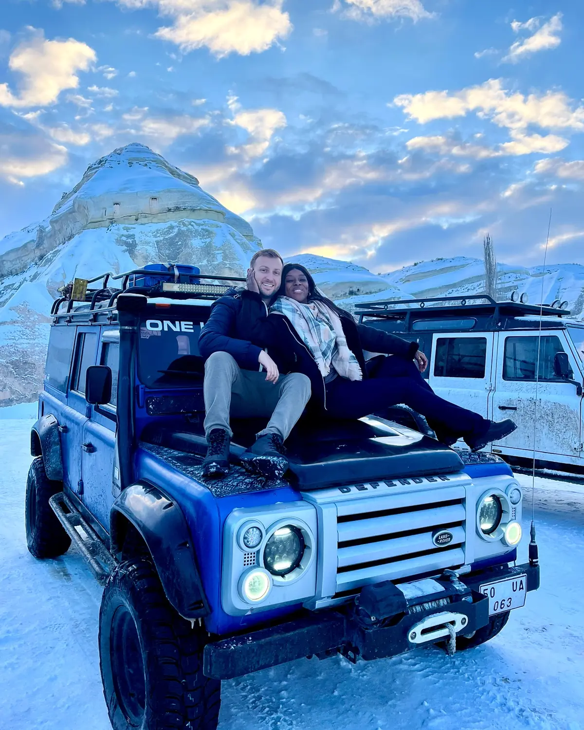 Us sitting on the hood of a blue Land Rover Defender at snowy sunrise among fairy chimneys