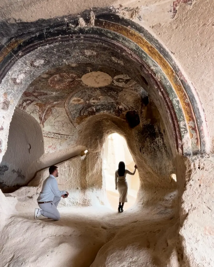 Andrew down on one knee proposing inside a frescoed rock-cut cave church as Melody walks through the arch