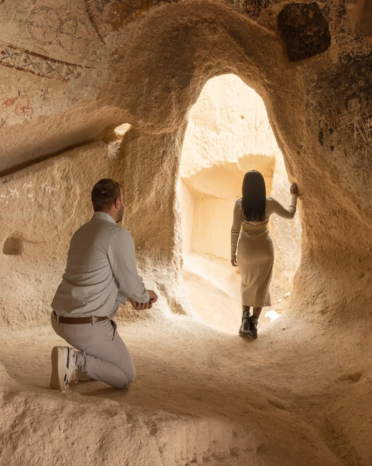 Andrew on one knee in a frescoed cave church as Melody stands in the glowing archway