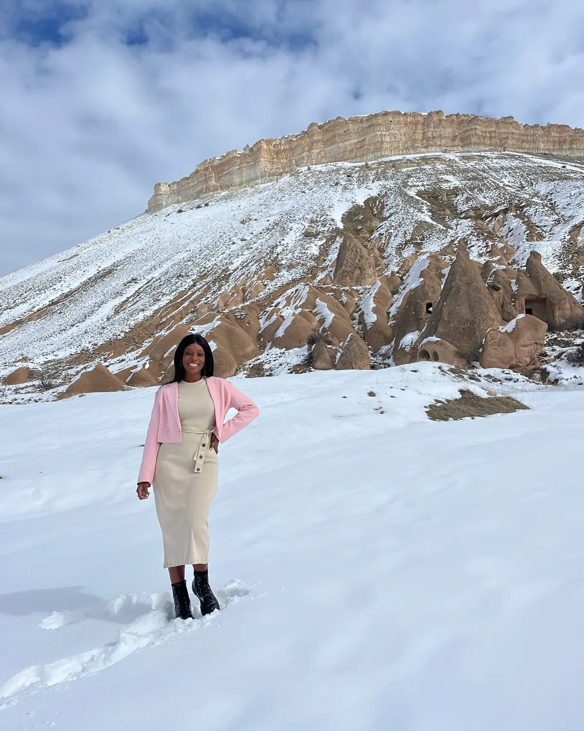 Melody in a pink jacket standing in the snow below a cliff of cave dwellings