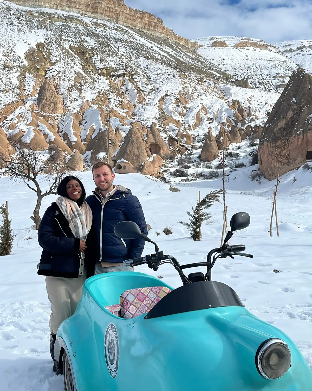 Us beside a turquoise sidecar trike in the snow with fairy chimneys and cliffs behind