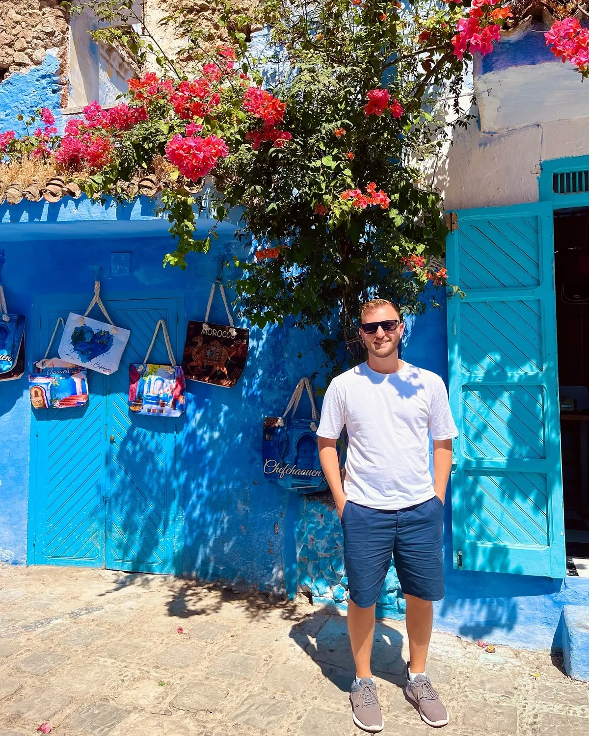 Andrew under a bougainvillea tree against a bright blue wall hung with souvenirs