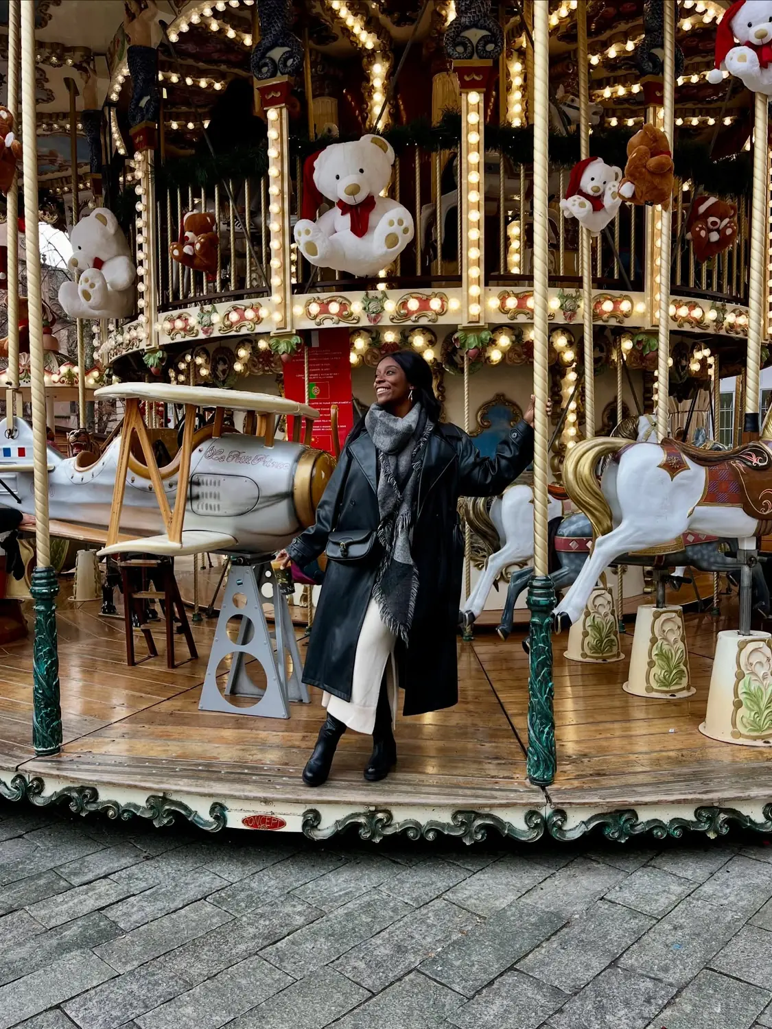 Melody beside a vintage carousel strung with lights and giant teddy bears in Colmar