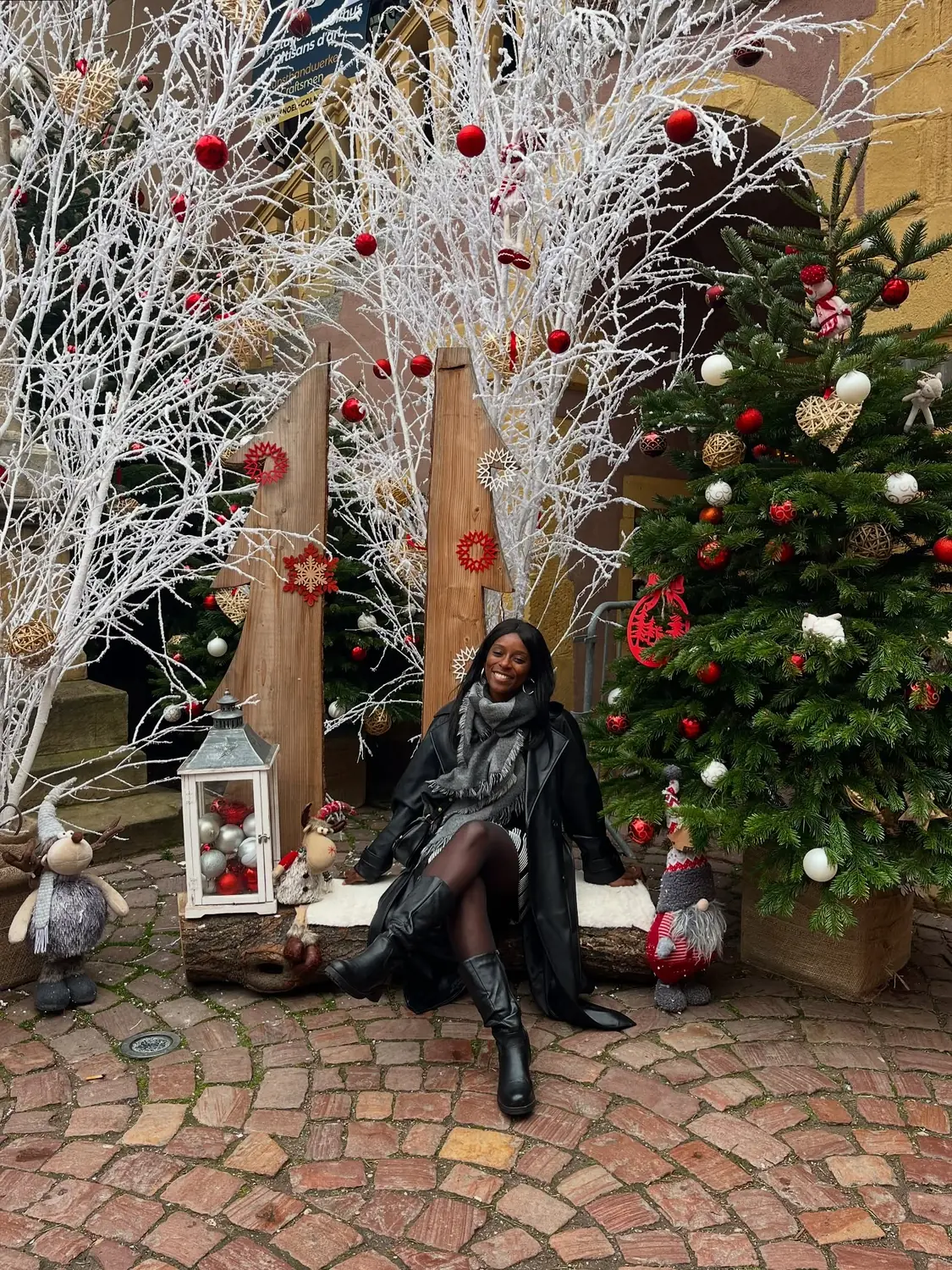 Melody seated in a festive Colmar courtyard photo spot with frosted white trees and a decorated Christmas tree