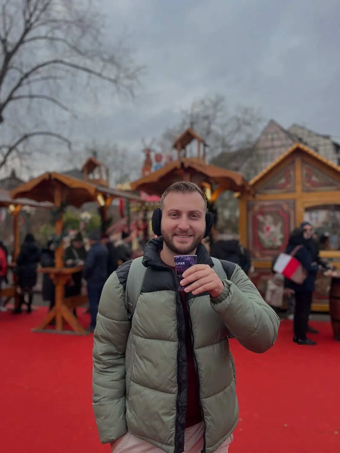 Andrew holding a purple Colmar mulled-wine cup at a Christmas market lined with wooden chalets