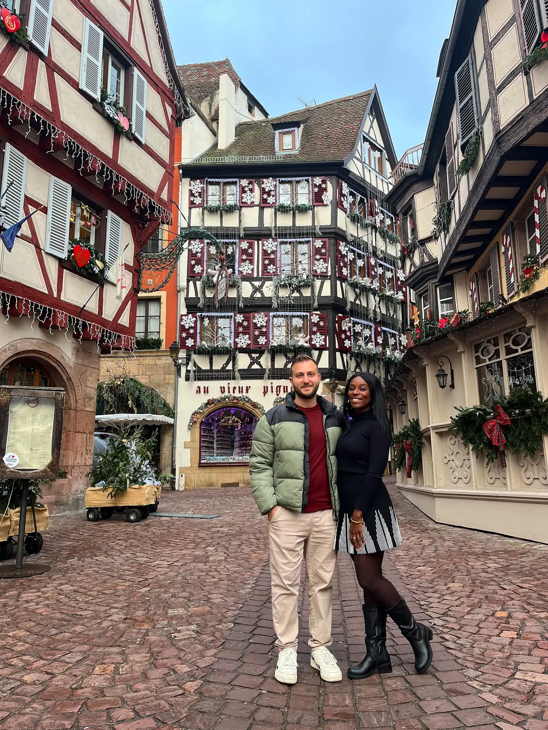 Us standing in the cobbled street beneath the snowflake-decorated Au Vieux Pignon facade in Colmar