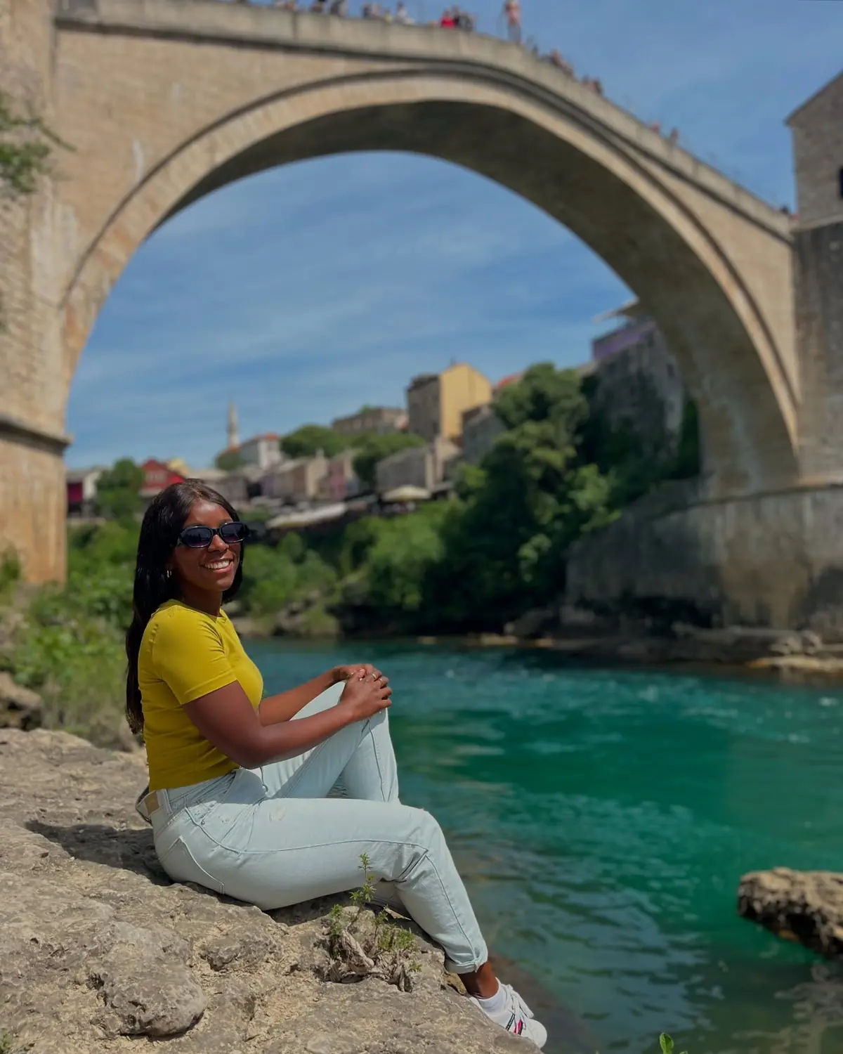 Melody sitting on the riverbank framed by the arch of Mostar's Old Bridge