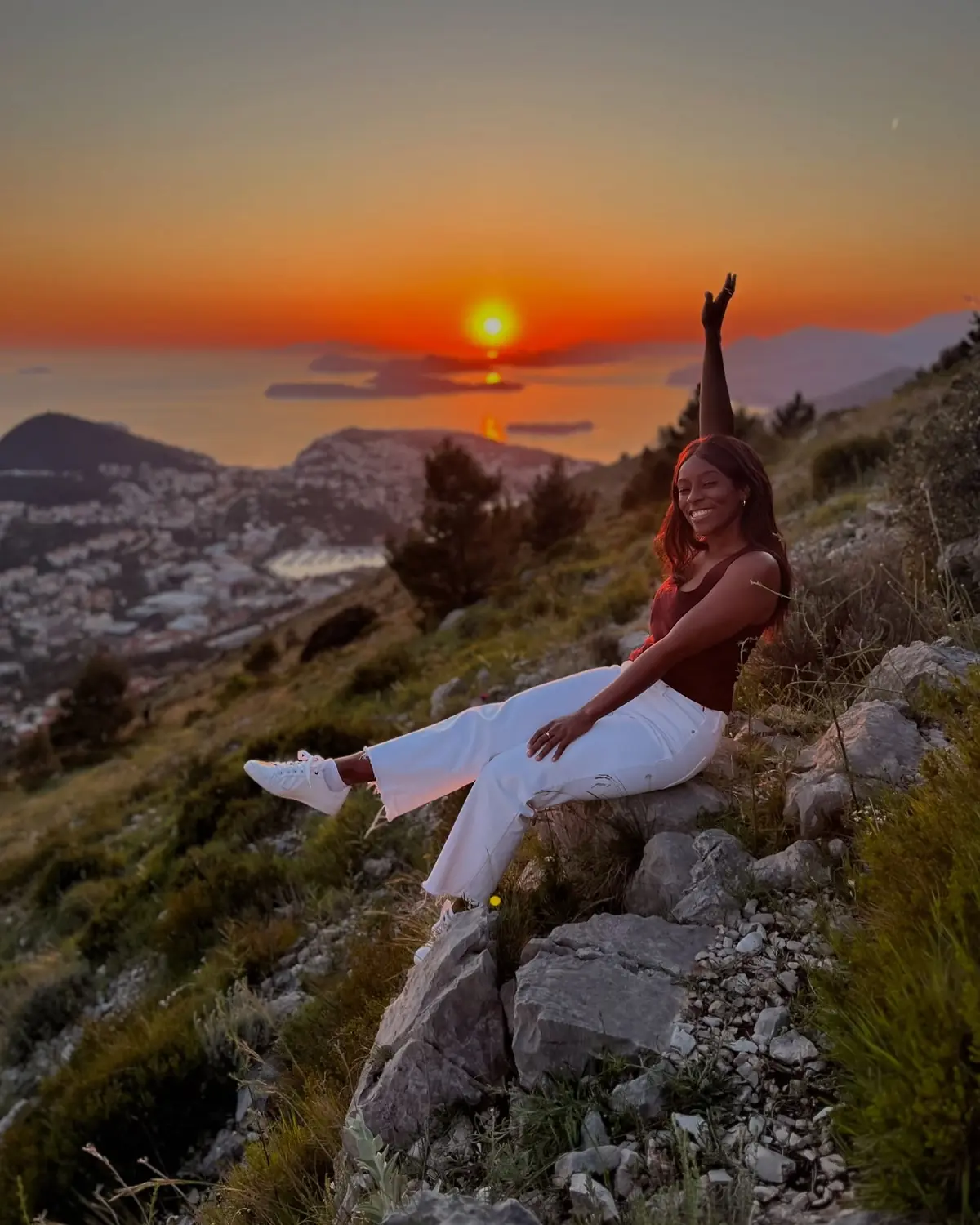 Melody on the mountainside with an arm raised as the sun sets over the islands below Dubrovnik