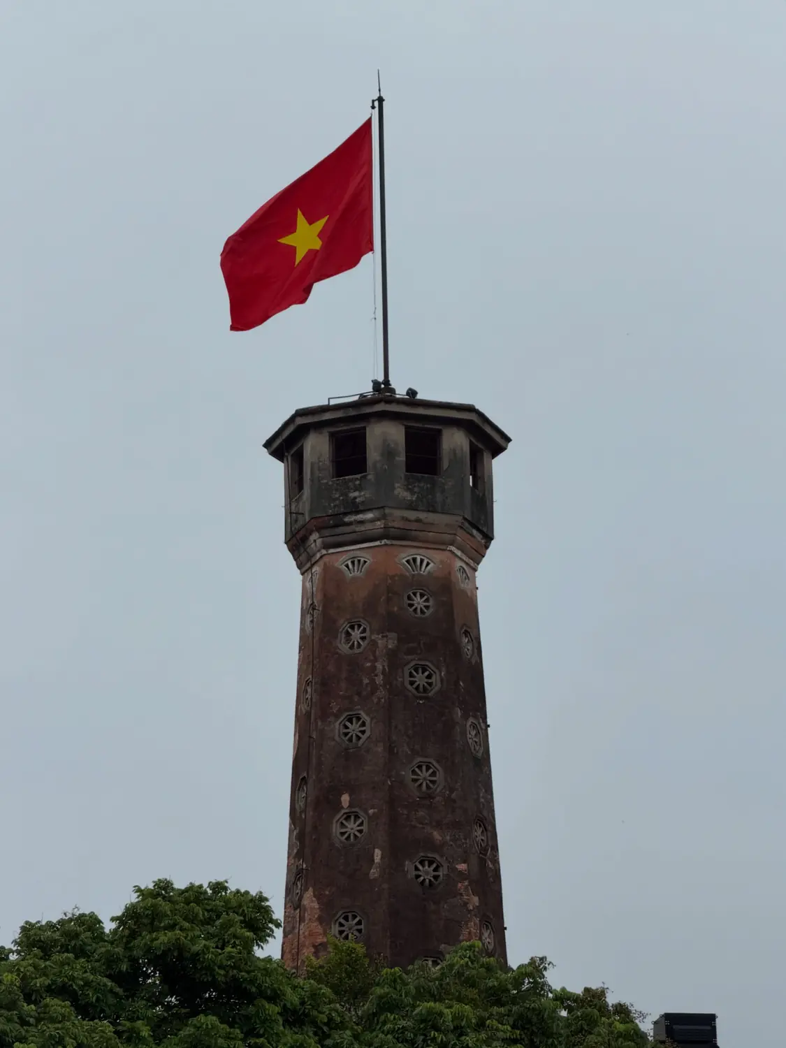 The Hanoi Flag Tower flying a large red Vietnamese flag against a grey sky