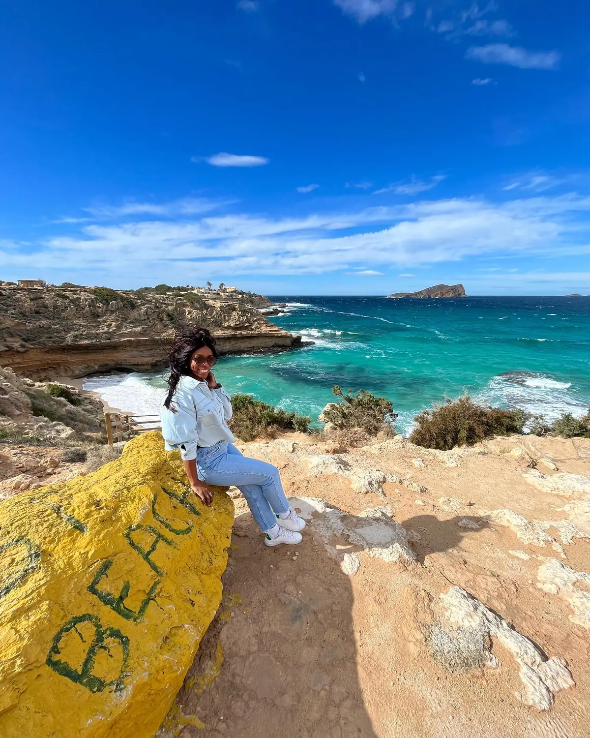Melody perched on a painted rock above a turquoise cove and a tiny island off Ibiza