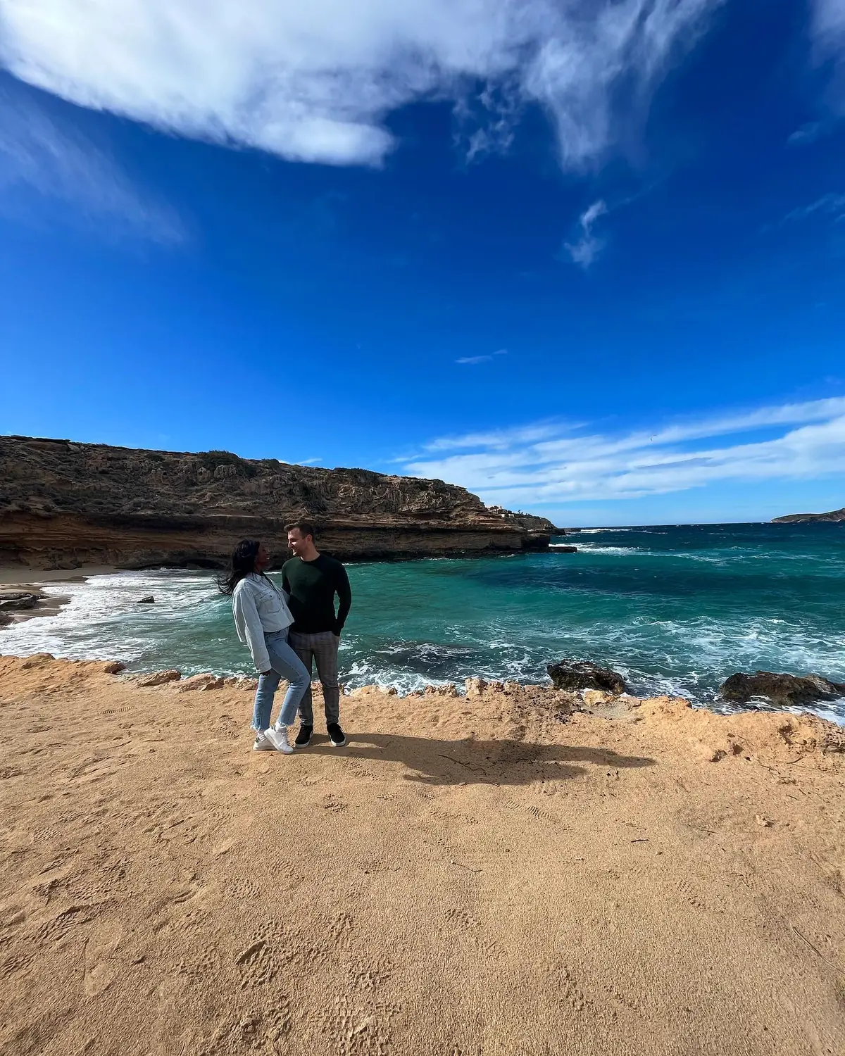 Us on the sand of a quiet Ibiza cove with cliffs and deep-blue water behind