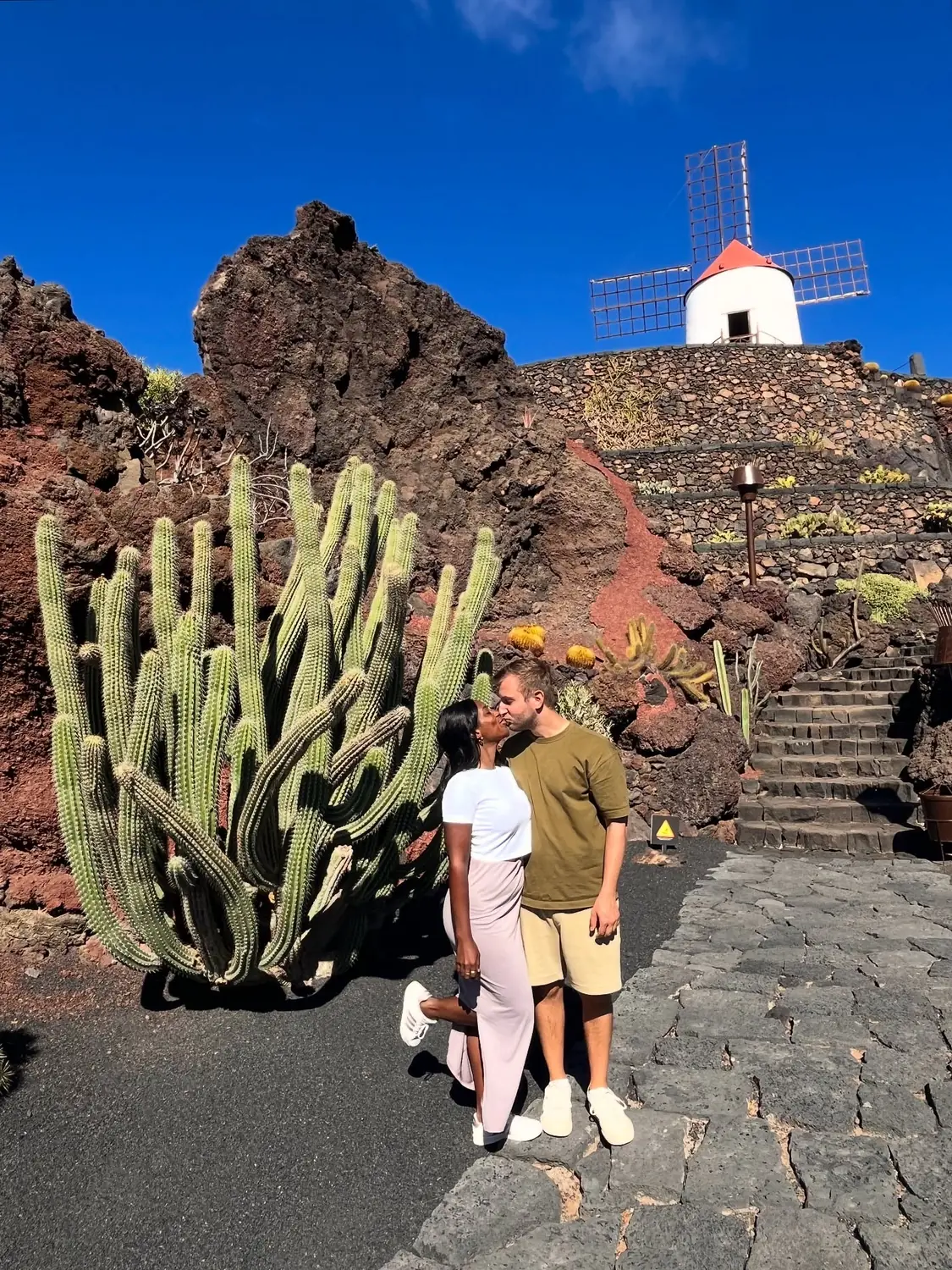 Us kissing beside a tall cactus with the cactus garden's windmill behind
