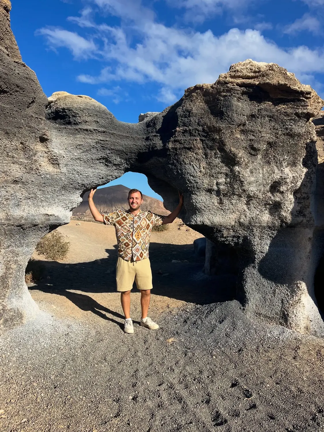 Andrew framed inside a natural volcanic rock arch with a cone-shaped volcano beyond