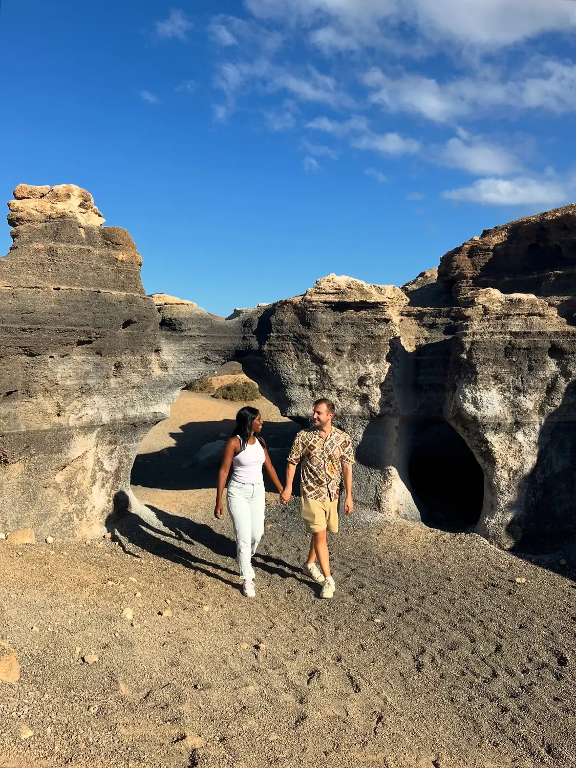 Us holding hands walking past pale eroded rock formations and a cave opening