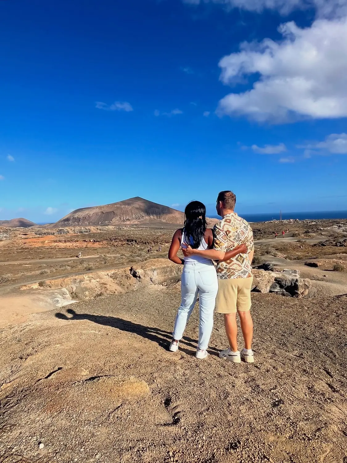 Us from behind looking out over a vast volcanic plain toward a large cone volcano and the sea
