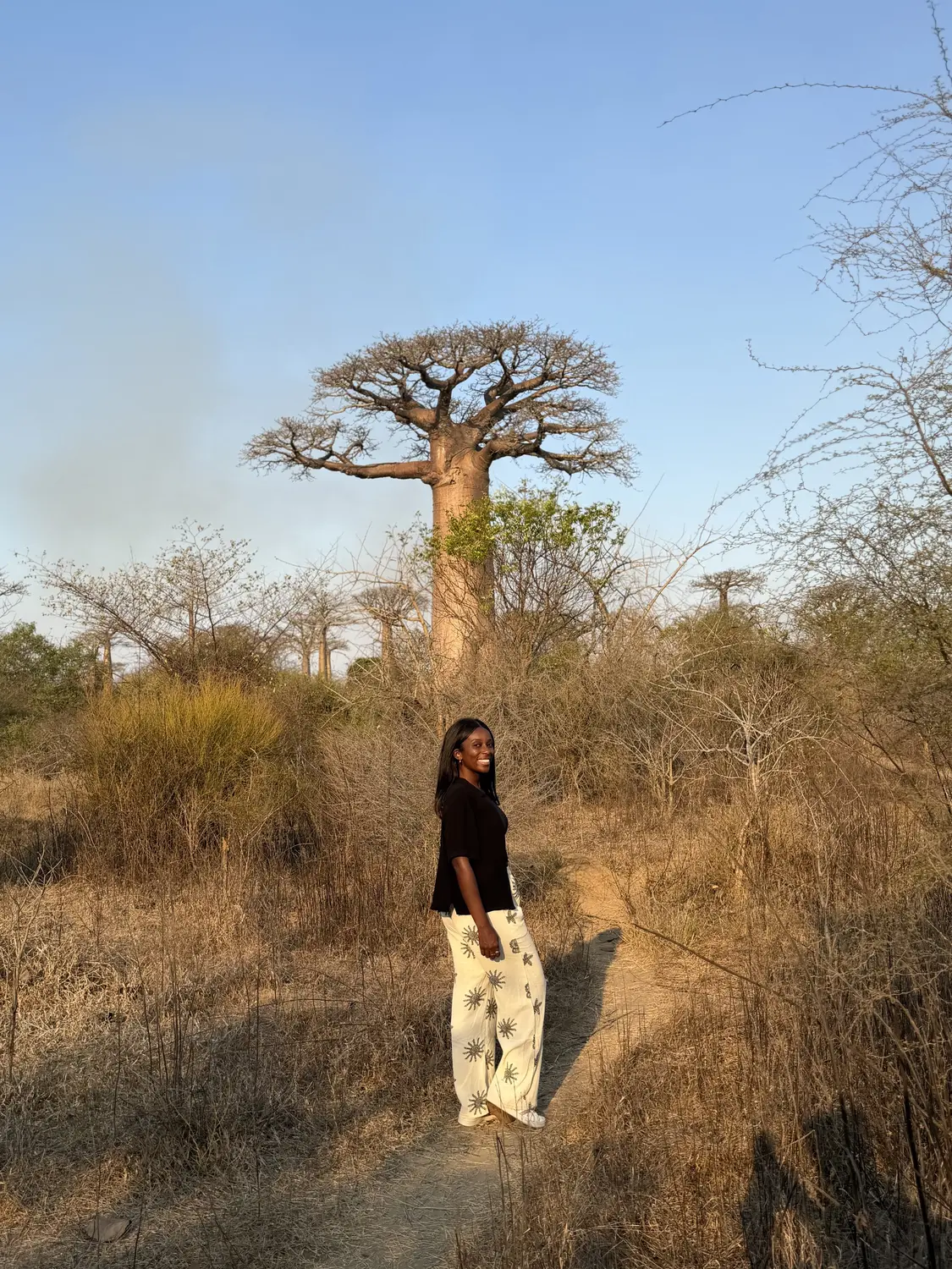Melody smiling on a dirt path in front of a huge baobab in warm evening light