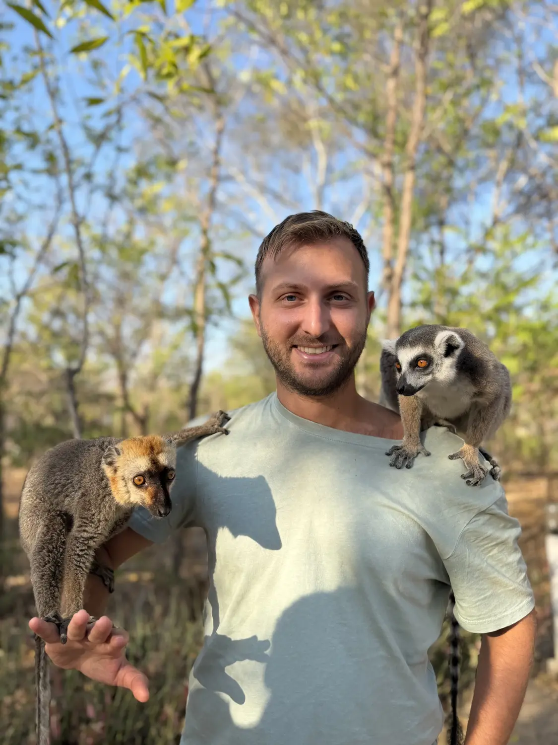 Andrew smiling with a ring-tailed lemur and a red-fronted lemur perched on his shoulders