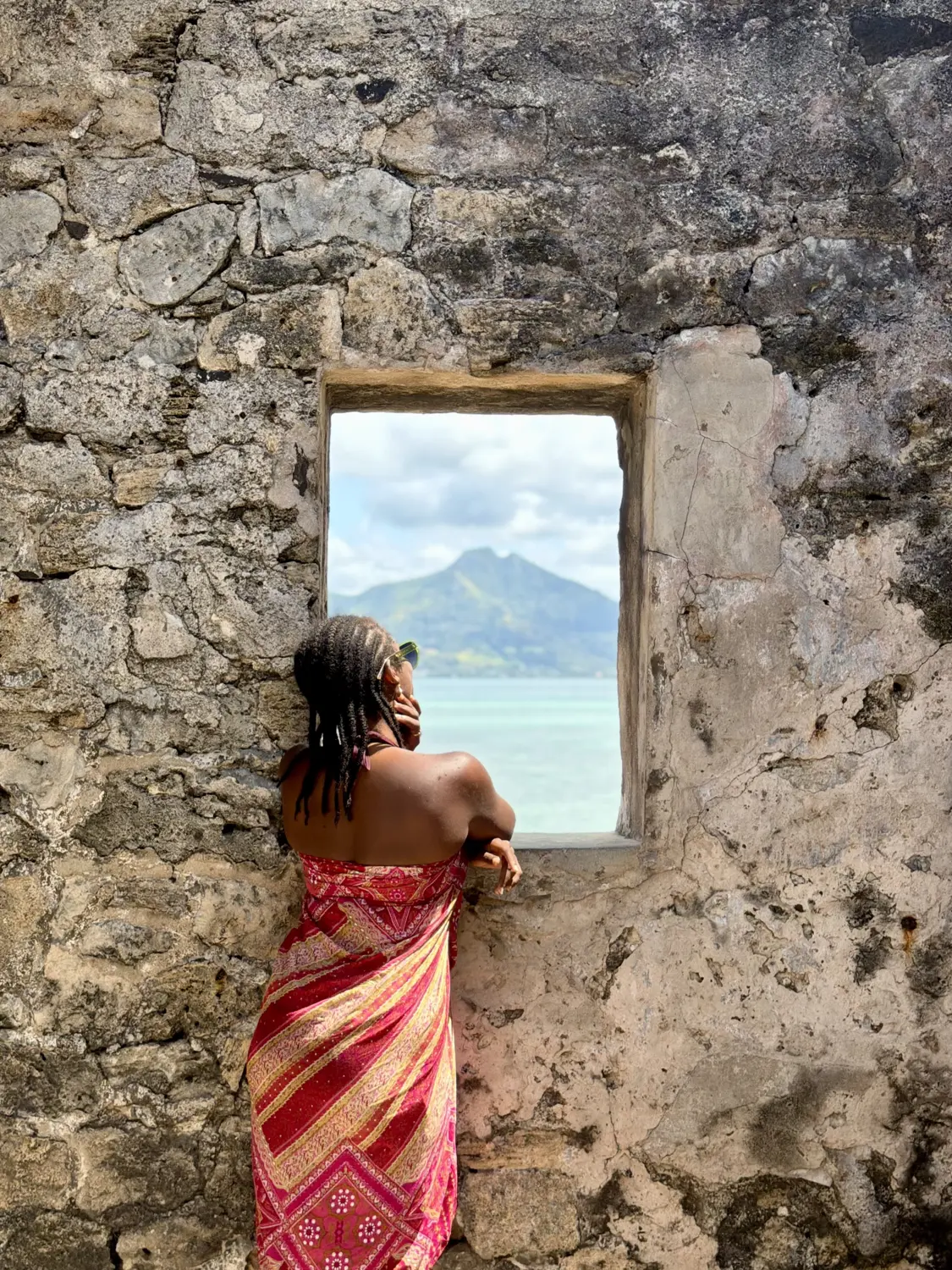 Melody looking out of a window in old stone ruins towards a green mountain and the sea