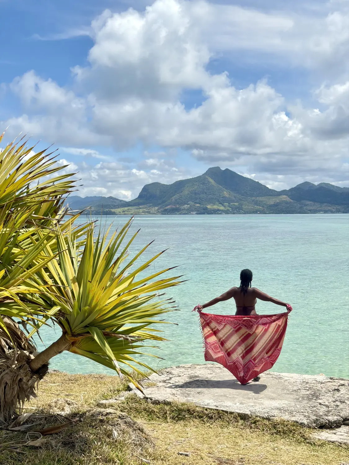 Melody holding out a pink sarong facing turquoise water and green mountains