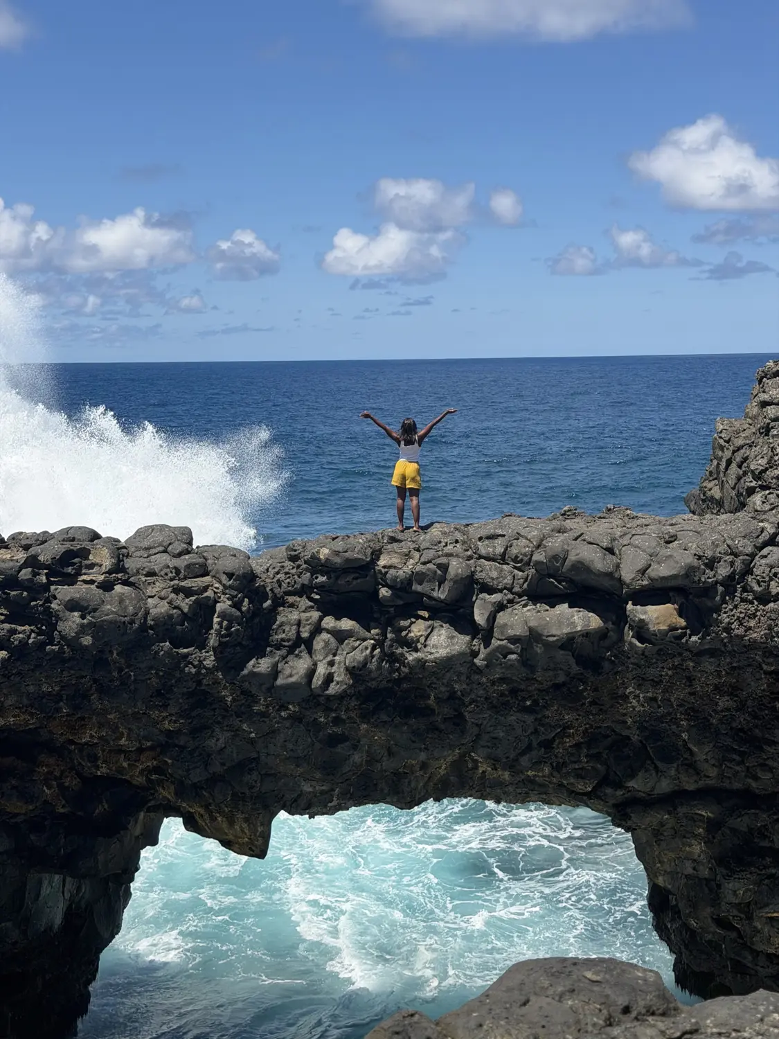Melody with her arms raised on top of a natural rock bridge with turquoise waves rushing underneath