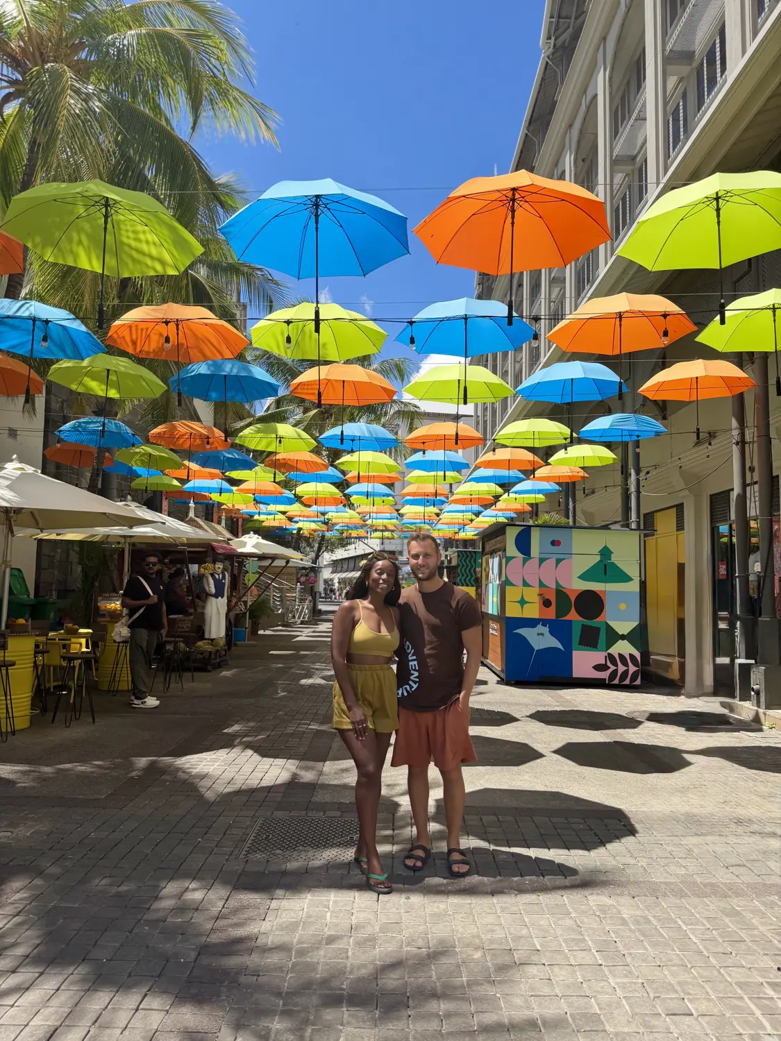 Us standing under a canopy of hanging blue, orange and green umbrellas in Port Louis