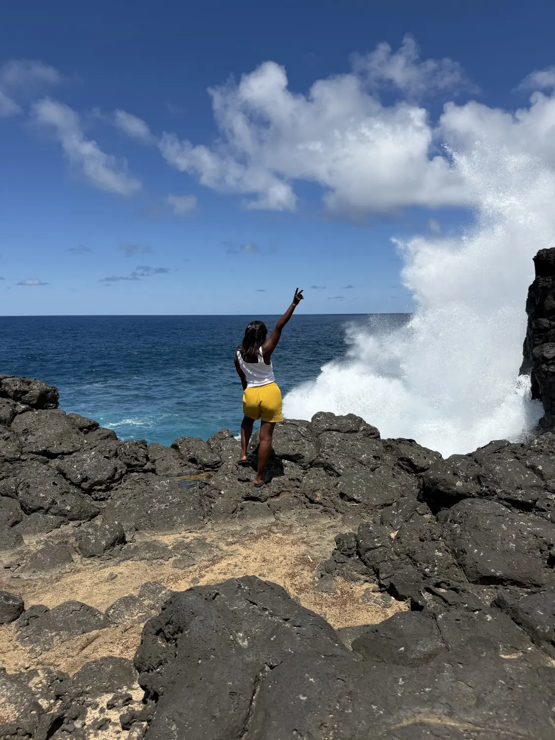 Melody giving a peace sign on black lava rocks as a huge wave explodes beside her