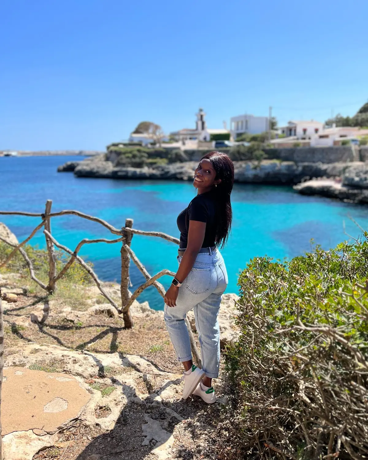 Melody at a cove viewpoint with a wooden fence, turquoise water and a lighthouse behind in Menorca