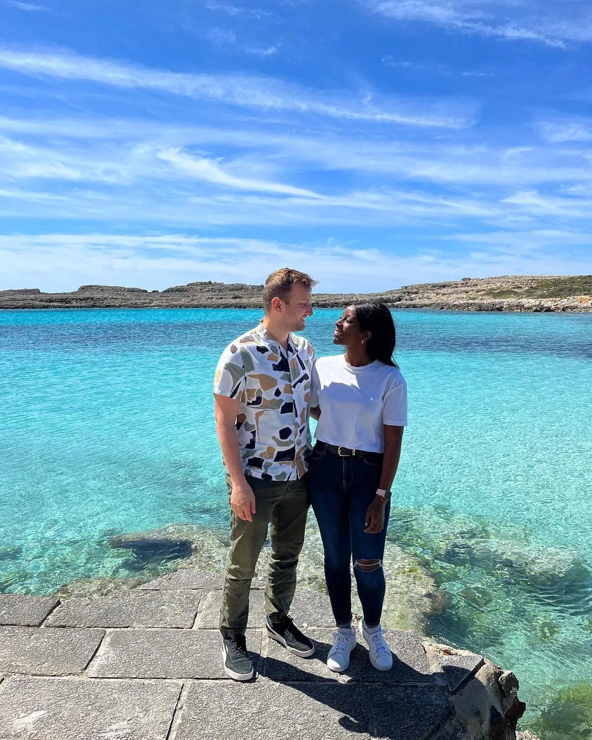 Us standing on stone beside impossibly clear turquoise water in Menorca, the Balearic Islands