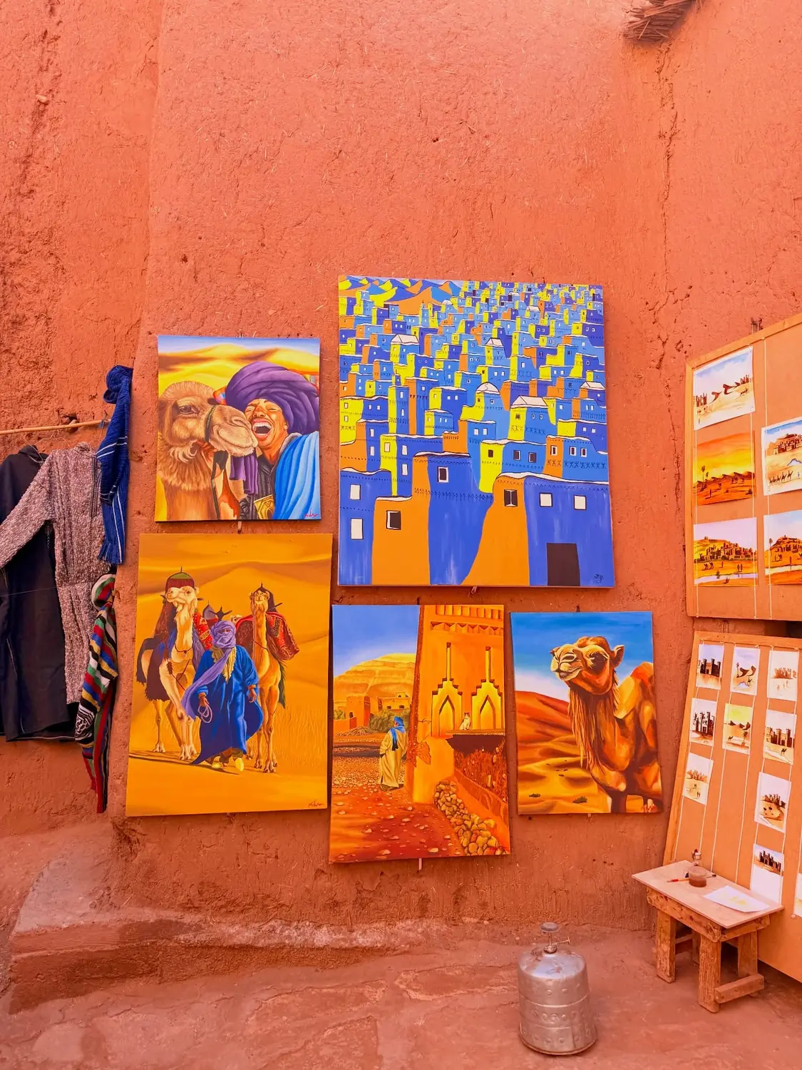 Brightly painted canvases of camels and kasbahs on a red mud wall at a roadside art stall