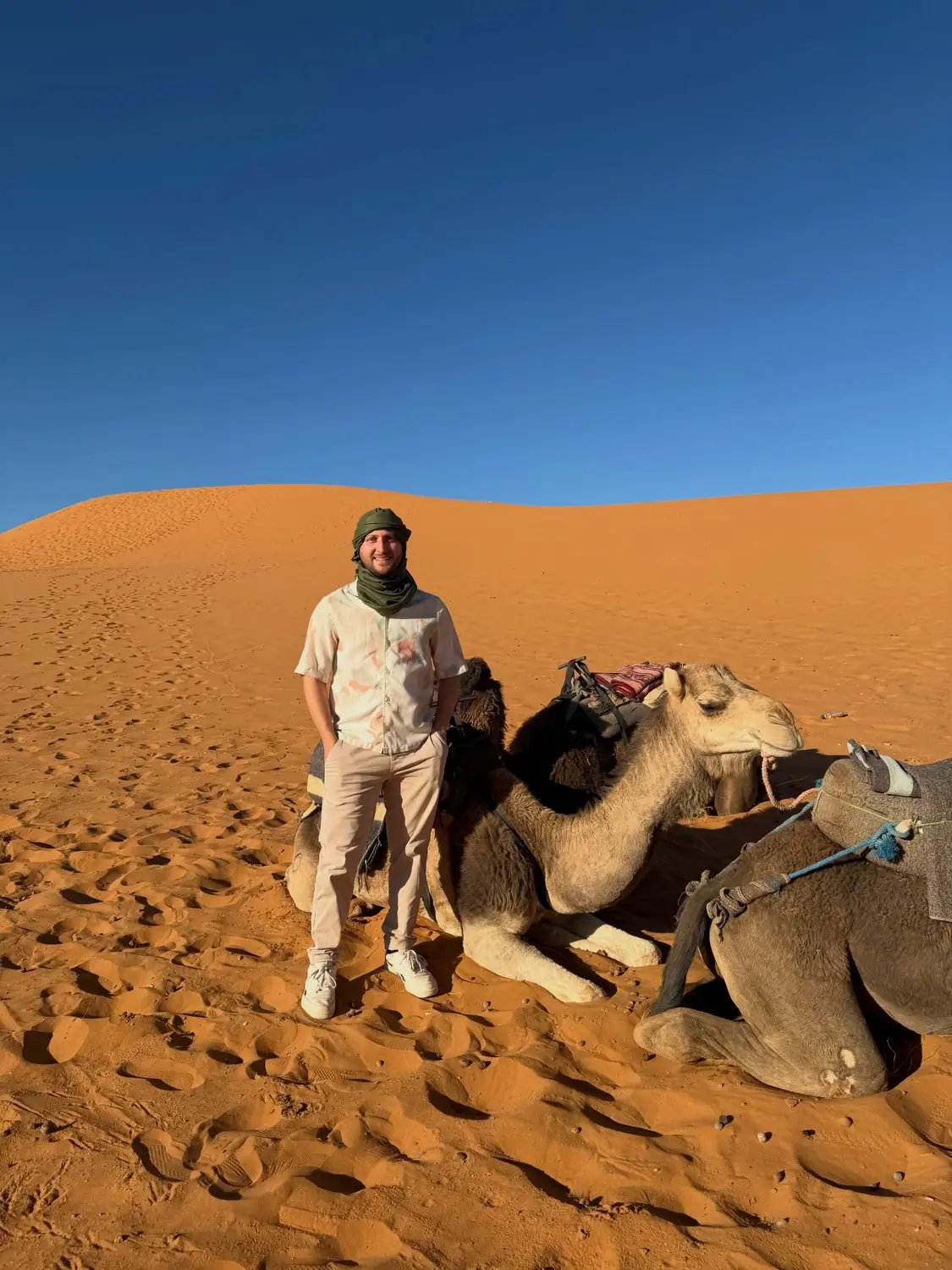 Andrew in a headscarf beside kneeling camels on the orange dunes at Merzouga