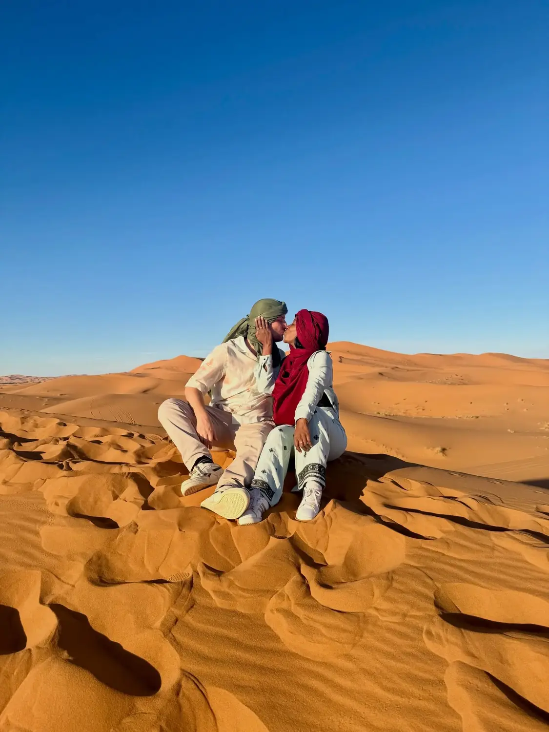 Us kissing on an orange Sahara dune in Berber headscarves under a clear blue sky at Merzouga