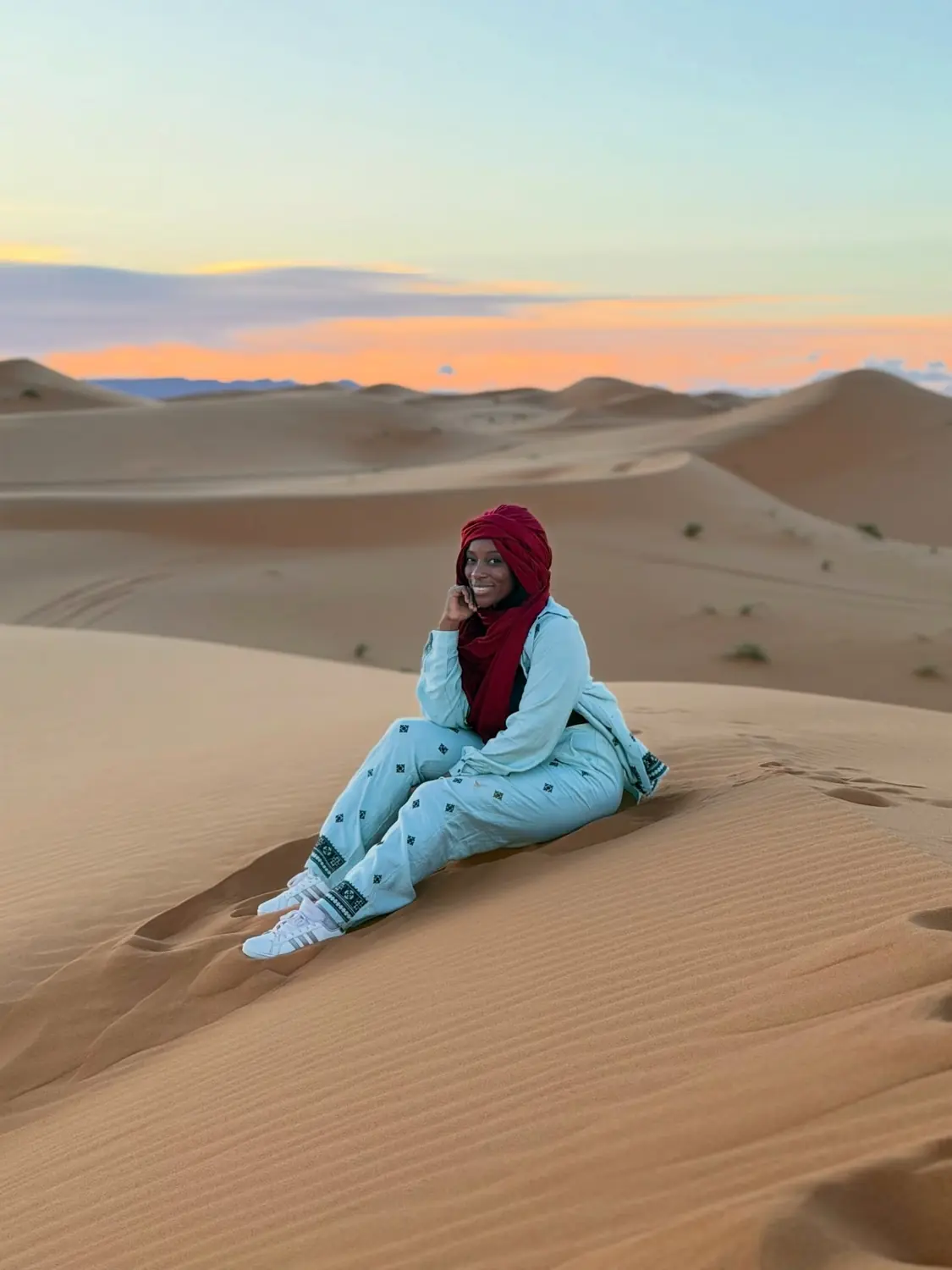Melody in a red headscarf sitting on a dune crest under a pastel dawn sky