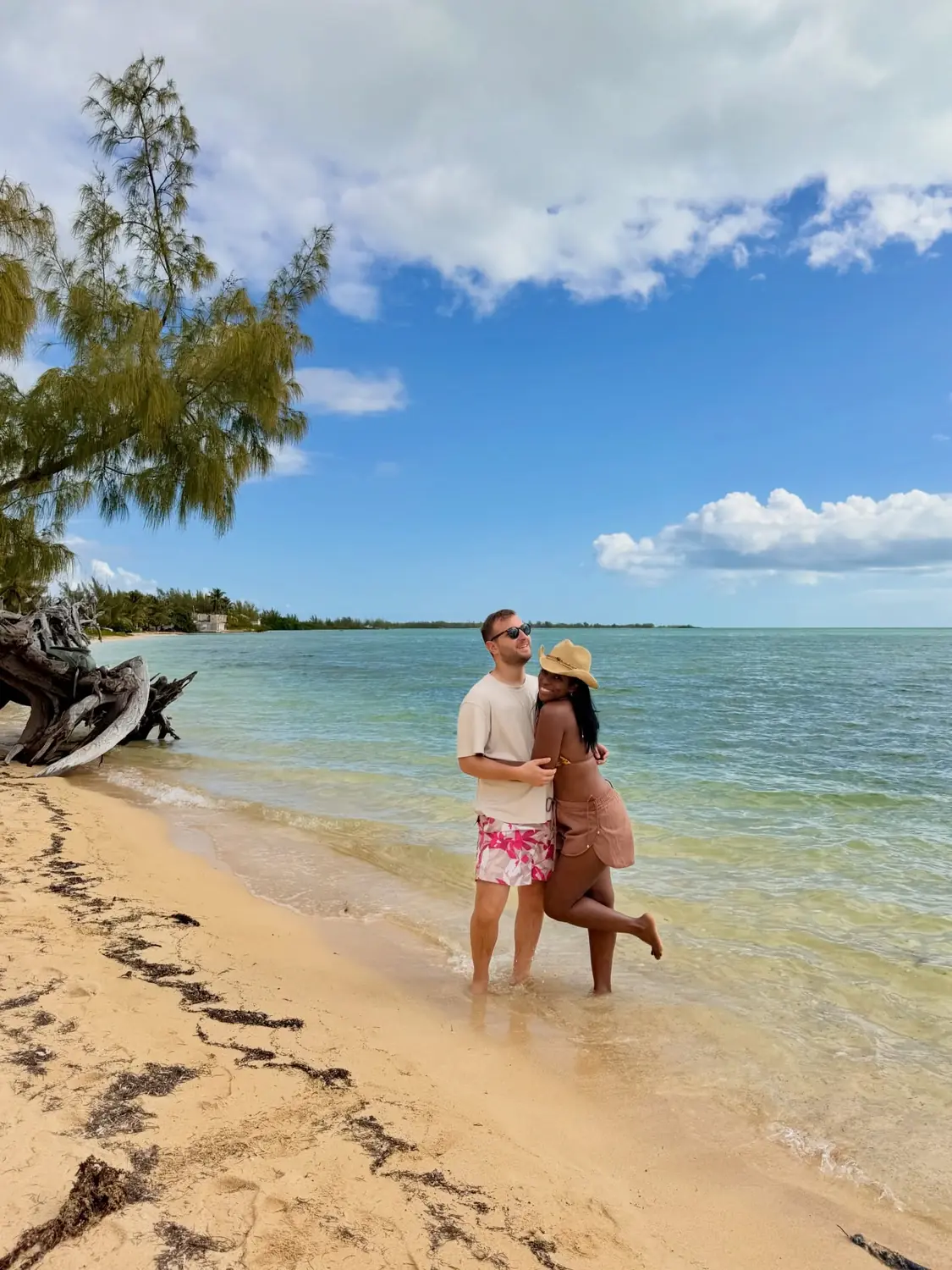 Us hugging on a Bahamas beach beneath a leaning palm and driftwood, turquoise water behind