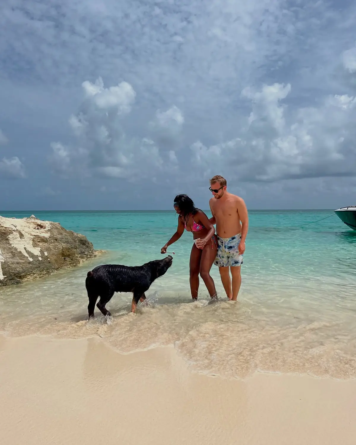 Us feeding a black swimming pig at the shoreline with a boat moored behind