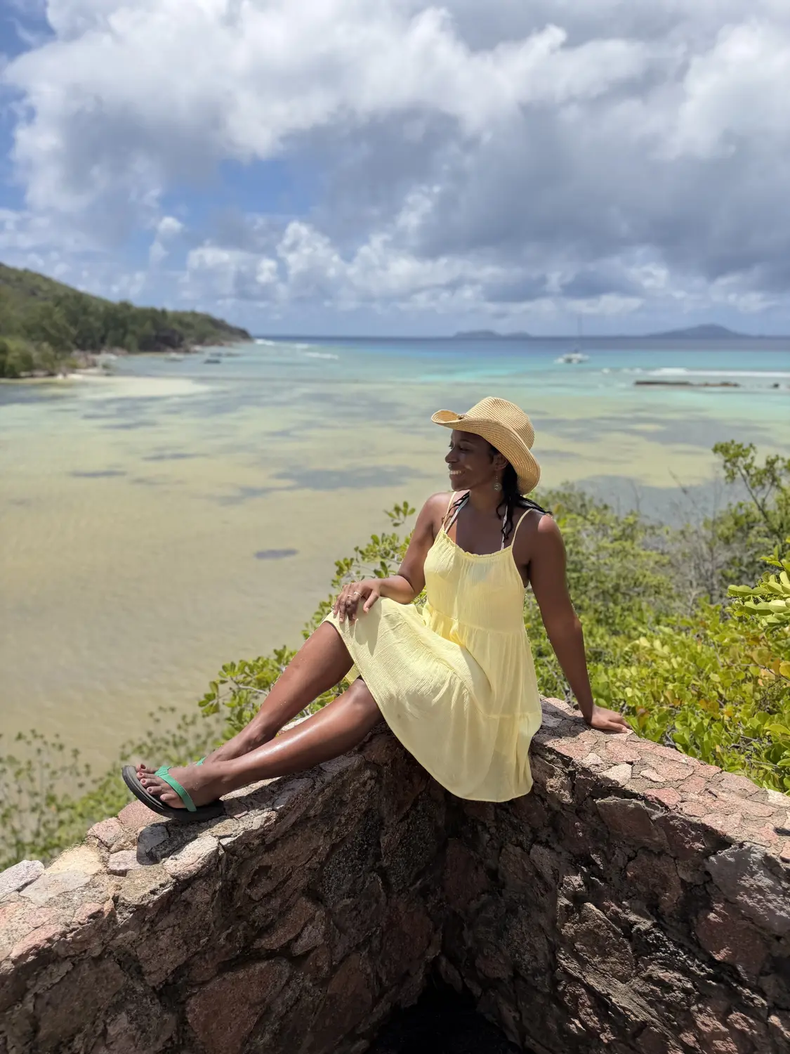 Melody sitting on a stone wall above a wide, shallow turquoise lagoon on Curieuse Island