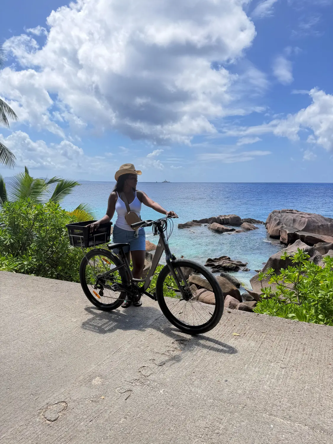 Melody with her rented bike on a coastal road above pink granite rocks and blue sea, La Digue