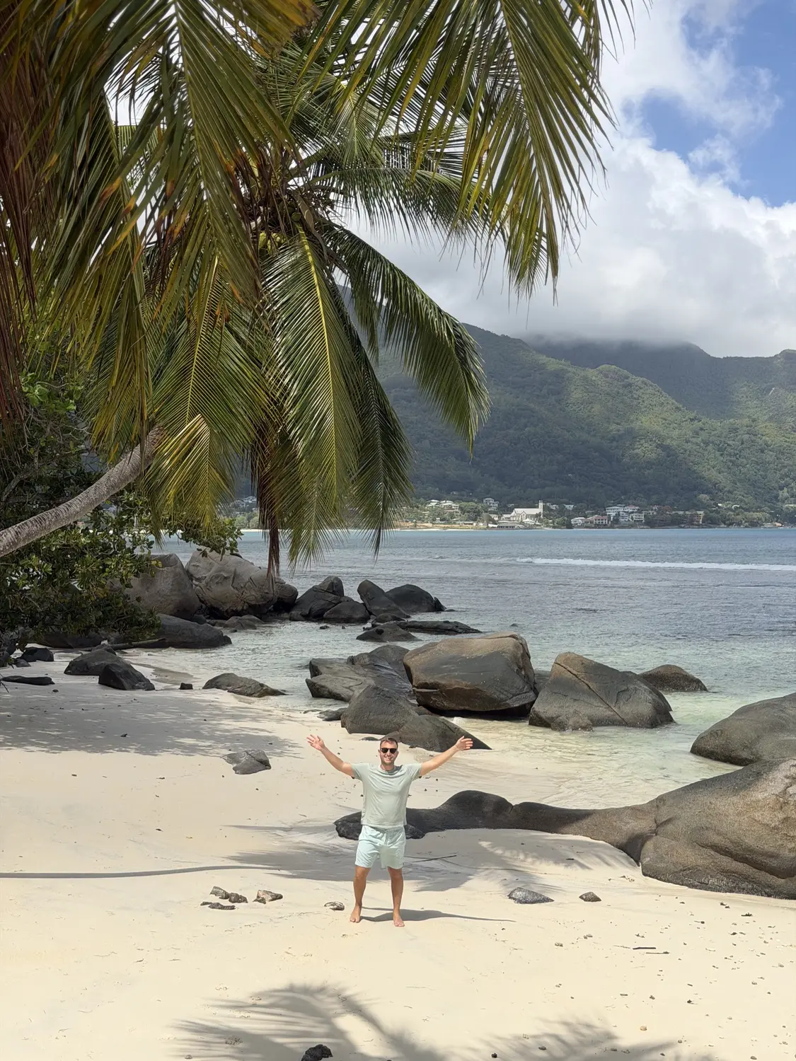 Andrew with his arms up on an empty cove under a palm tree, looking back across the bay to Beau Vallon, Mahé