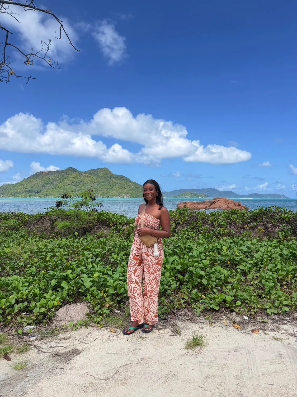 Melody standing on Praslin's coast with green islands and a red granite rock in the bay behind
