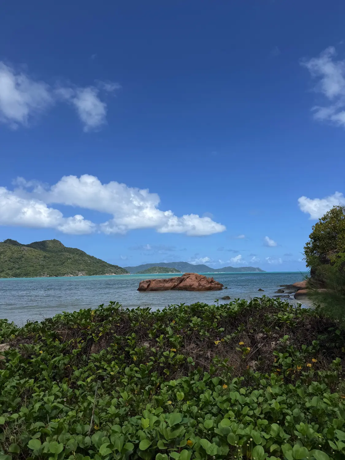 A red granite rock in turquoise water with green islands on the horizon, Praslin