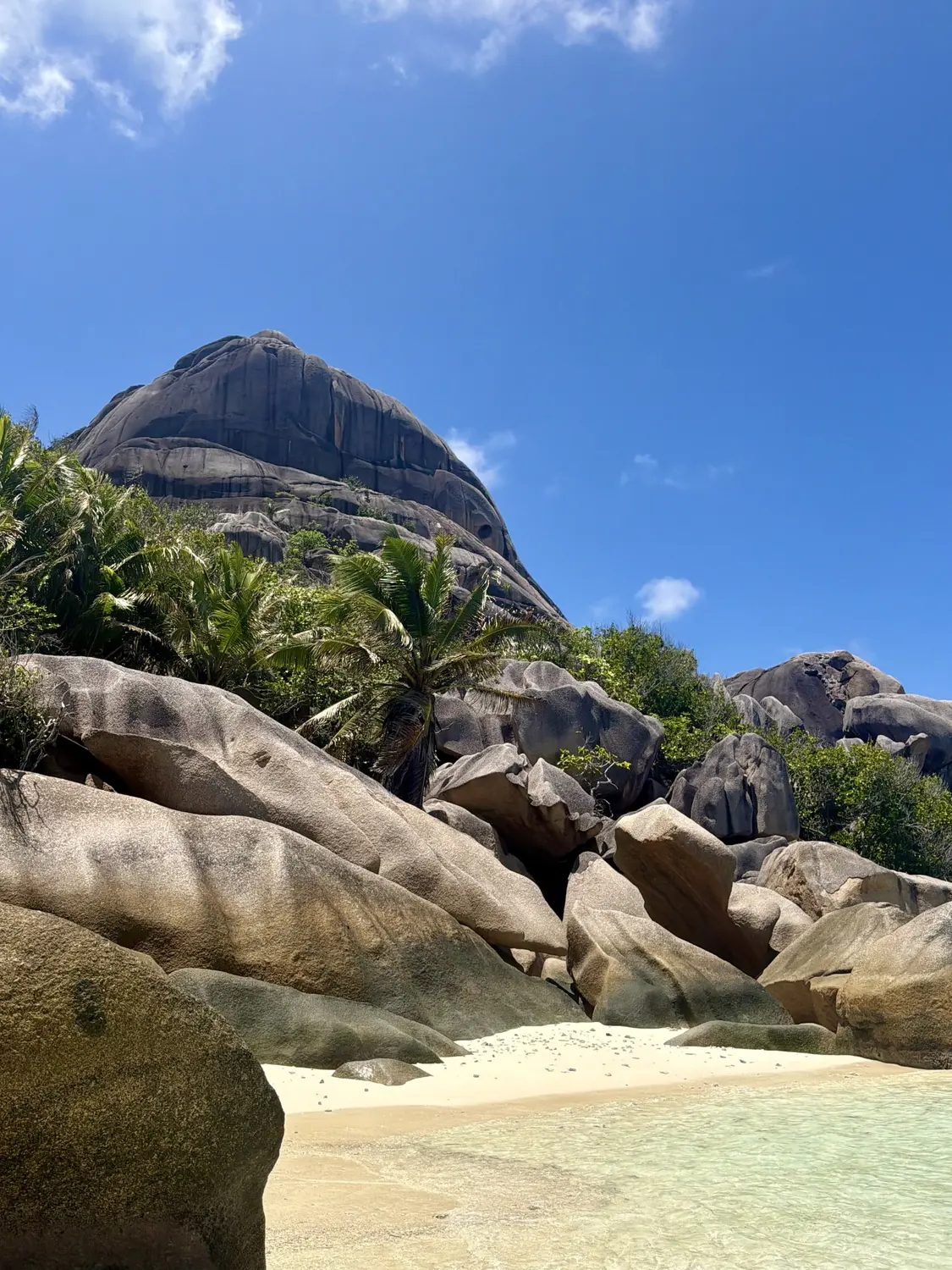 A huge granite dome rising above palms and boulders over a quiet white-sand cove, La Digue