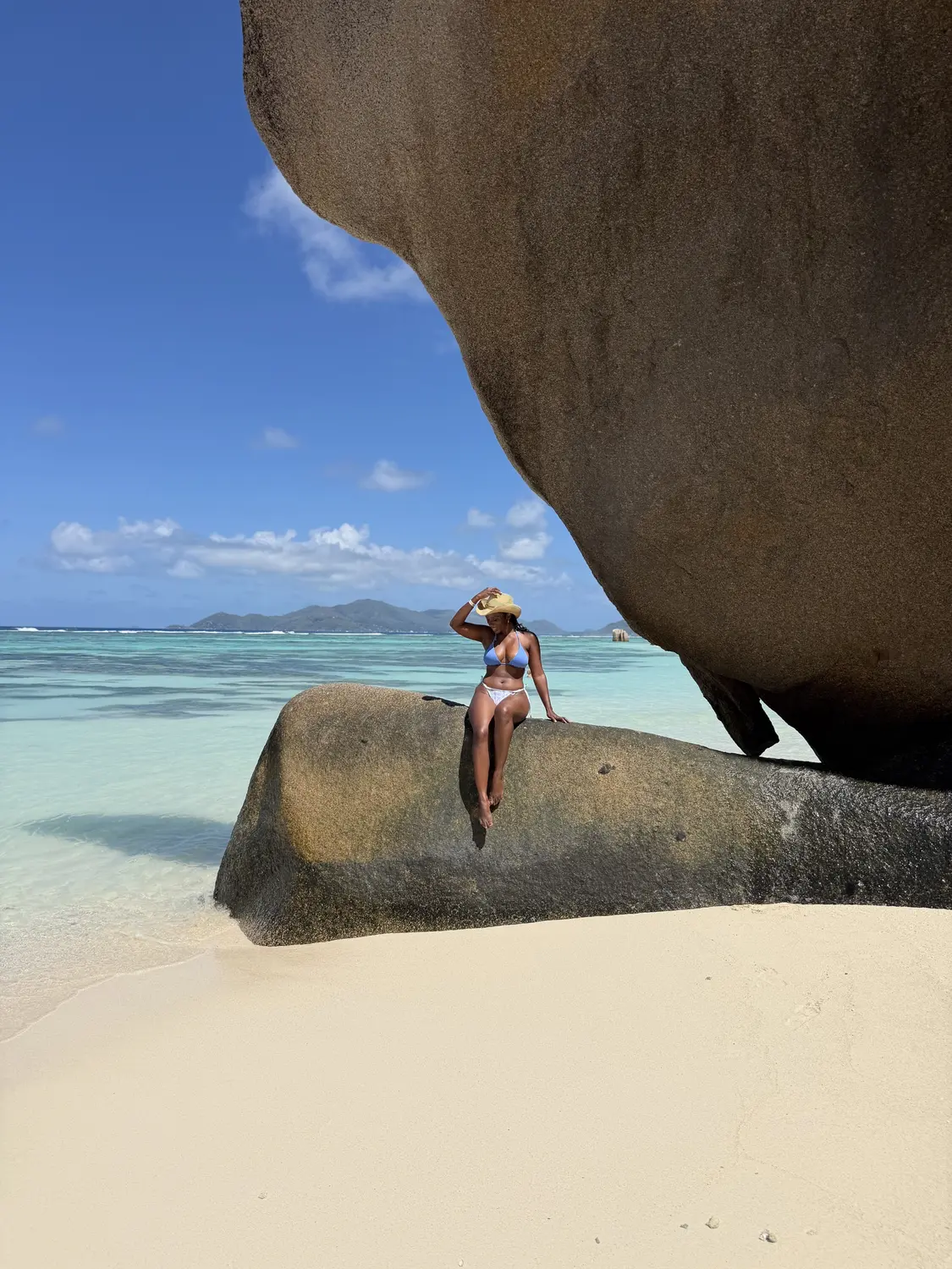 Melody holding her hat on a granite boulder beside the turquoise lagoon at Anse Source d'Argent