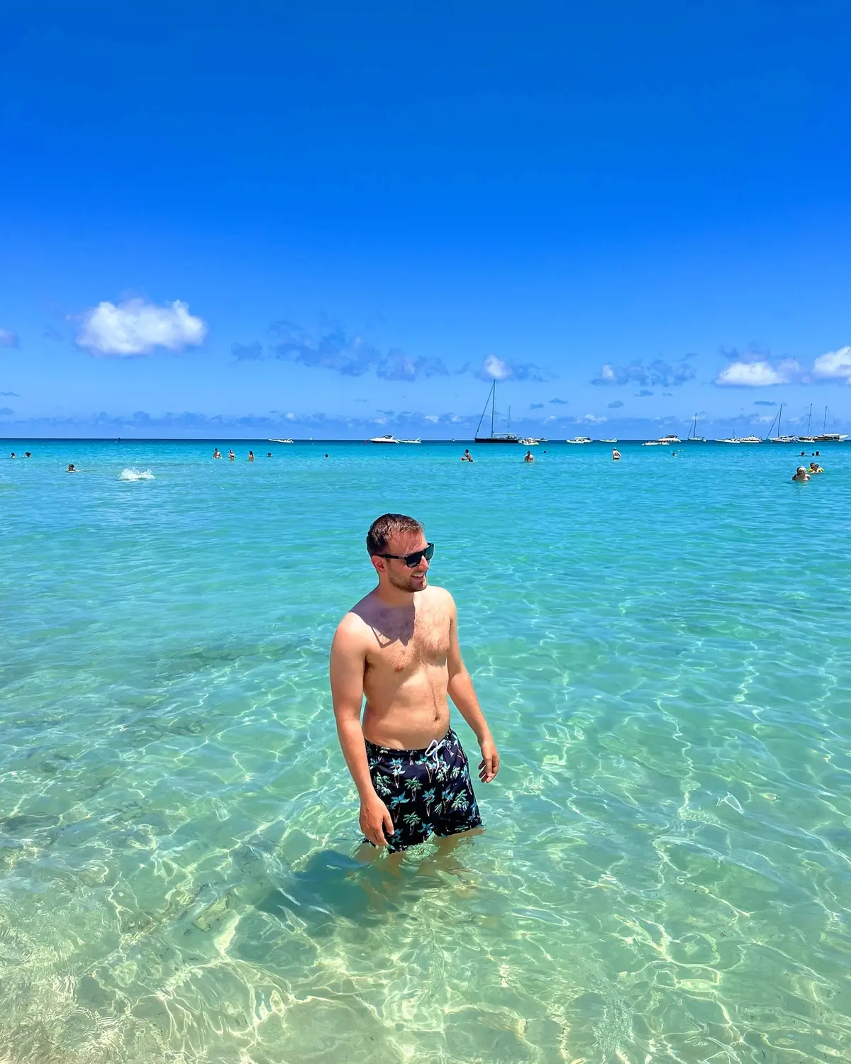 Andrew standing in the crystal-clear water at Mondello with boats behind