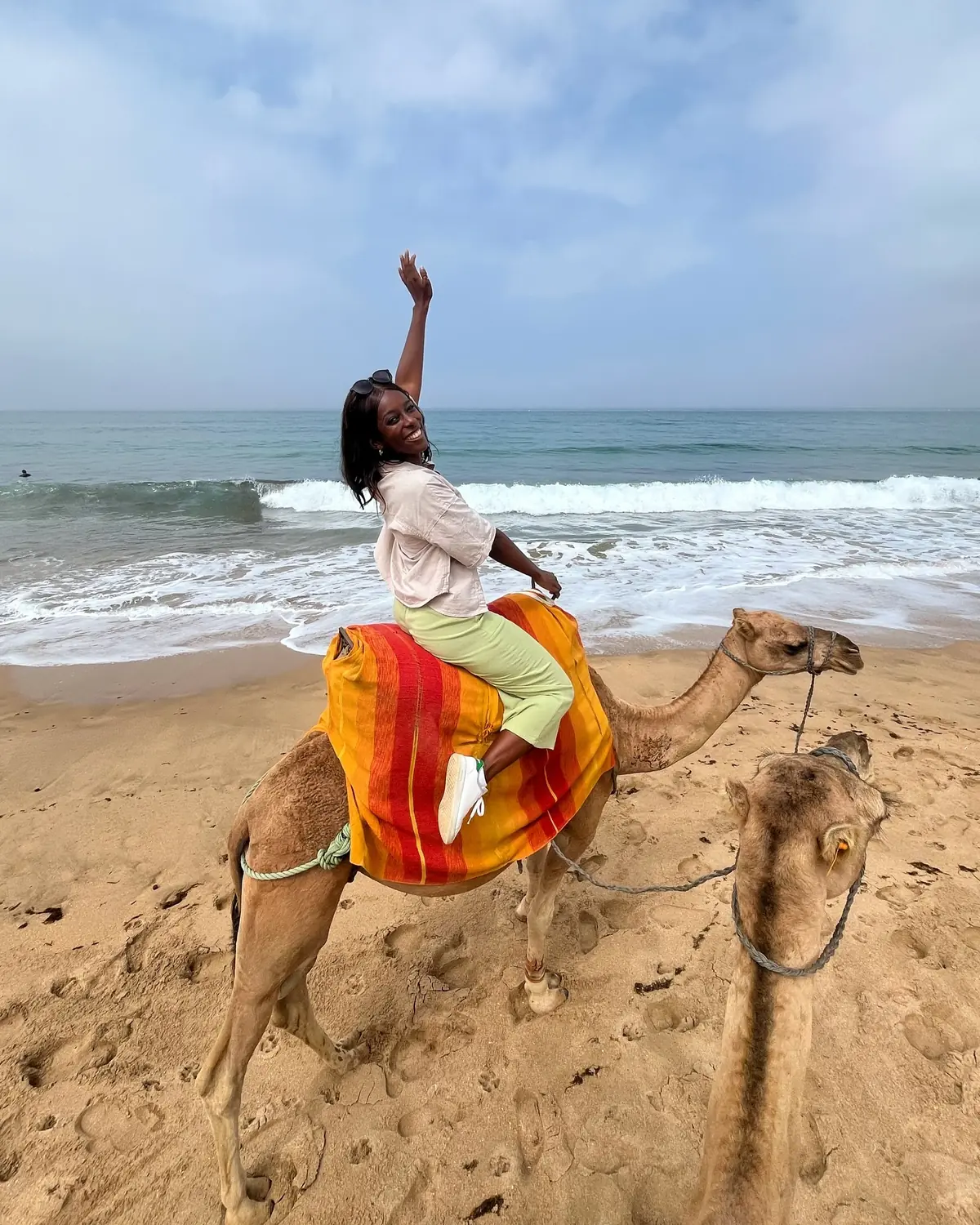 Melody on a camel by the shoreline, one arm raised, laughing