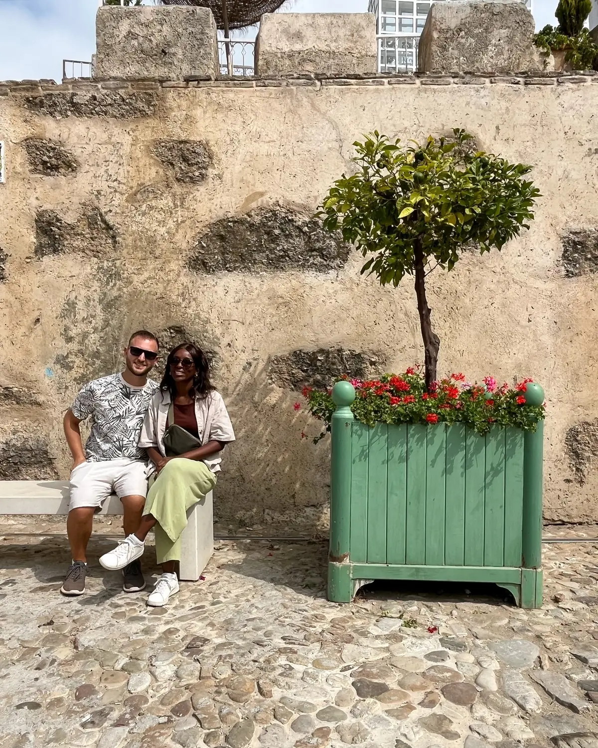 Us on a stone bench beside a potted orange tree with red geraniums in the old town
