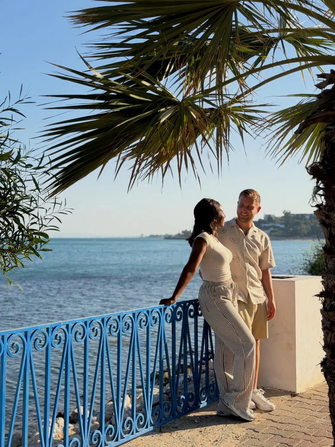 Us on a palm-shaded seafront promenade with a blue railing at golden hour