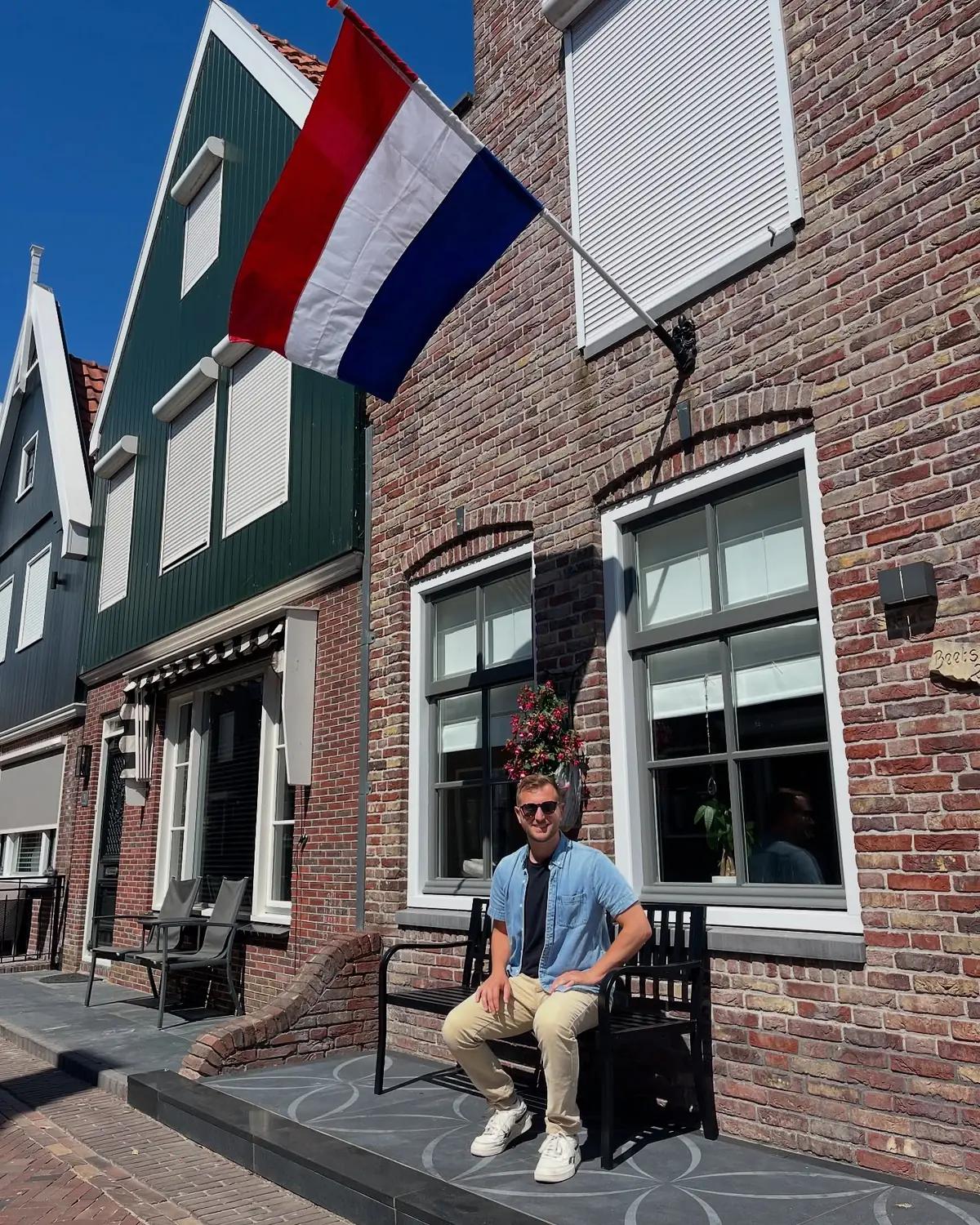 Andrew on a bench beneath a Dutch flag on a brick-house street in a fishing village